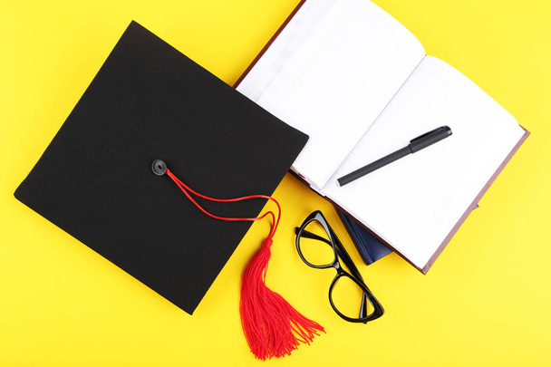Graduation cap with notepad, pen and glasses on yellow backgroun - Foto, Bild