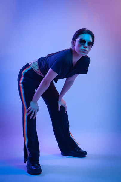 Mujer modelo de alta moda en camisetas de moda y pantalones con rayas en colores lgbt en luces de colores brillantes de neón azul y rosa, posando en el estudio. Concepto de moda y cultura Zine
 - Foto, Imagen
