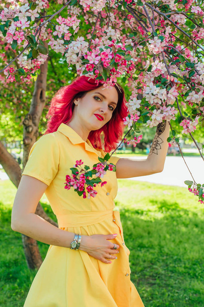 Beautiful and young woman with bright red hair and ref lips standing next to a blooming apple tree in a yellow dress. - 写真・画像