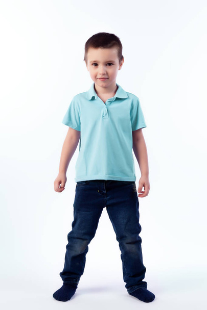 Little smiling boy with dark hair in blue jeans, blue polo t-shirt posing, laughing happily on a white isolated background in a photo studio. Portrait of happy joyful beautiful boy - Photo, Image