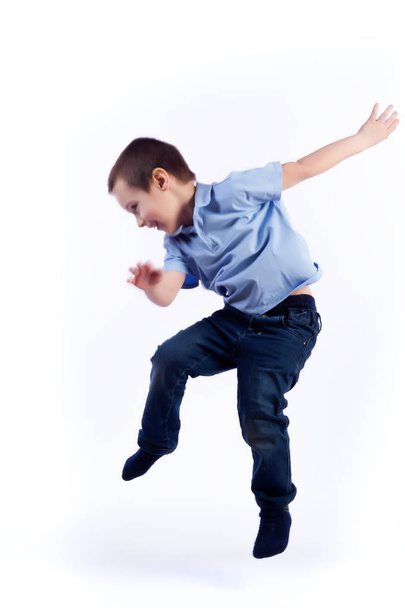 Little smiling boy with dark hair in blue jeans, blue polo t-shirt is jumping and having fun on an isolated white background in a photo studio - 写真・画像