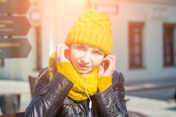 Portrait of a very attractive woman in a knitted hat and scarf with headphones in the ears on a city street - Фото, зображення