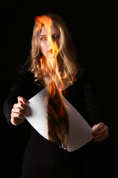 Beautiful young woman burning sheet of paper on dark background - Photo, Image