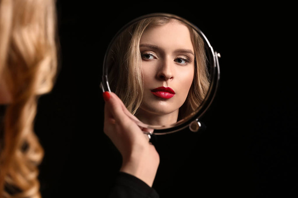Красива молода жінка з яскравим макіяжем дивиться у дзеркало на темному тлі
 - Фото, зображення