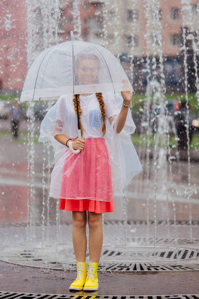 Menina bonita nova com duas tranças em botas amarelas e com guarda-chuva transparente fica perto da fonte
. - Foto, Imagem