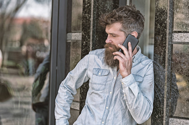 Man with beard and mustache on strict face talking, building on background. Bearded man speaking on cell phone. Communication concept. Hipster with beard speak on mobile phone outdoor - Foto, afbeelding