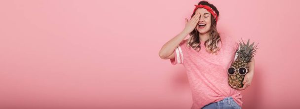 Menina bonita em camiseta rosa, fecha um olho, segura abacaxi
 - Foto, Imagem