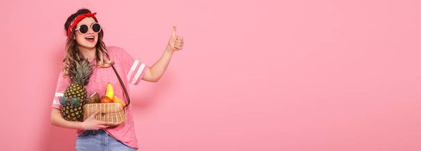 schönes junges Mädchen in rosa T-Shirt und Brille, hält einen vollen Strohsack mit Früchten auf rosa Hintergrund. Zeigt Fingerklasse - Foto, Bild