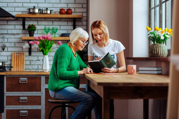 Young church volunteer reading Bible with pensioner at home kitchen - Photo, Image