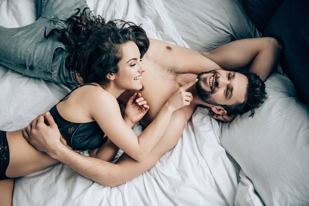overhead view of happy bearded man lying with smiling brunette woman in black underwear on bed - Photo, image