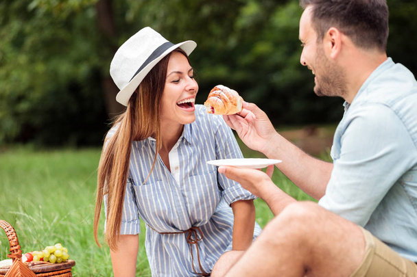 Feliz pareja joven tomando un desayuno relajante en el parque, comiendo croissants y tomando café
 - Foto, imagen