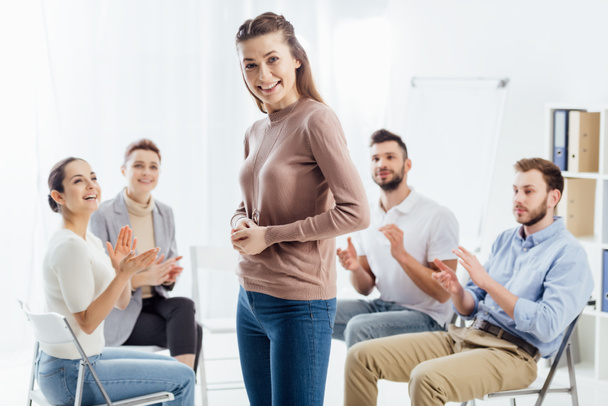 smiling woman looking at camera while people sitting and applauding during group therapy session - Photo, Image