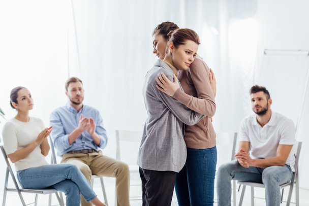 Frauen umarmen sich, während eine Gruppe von Menschen sitzt und während der Therapiesitzung applaudiert - Foto, Bild