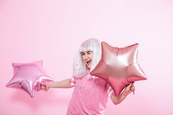 Αστείο κορίτσι με ασημένια μαλλιά δίνει ένα χαμόγελο και συγκίνηση σε ροζ φόντο. Νεαρή γυναίκα ή έφηβος κορίτσι με μπαλόνια και κομφετί - Φωτογραφία, εικόνα