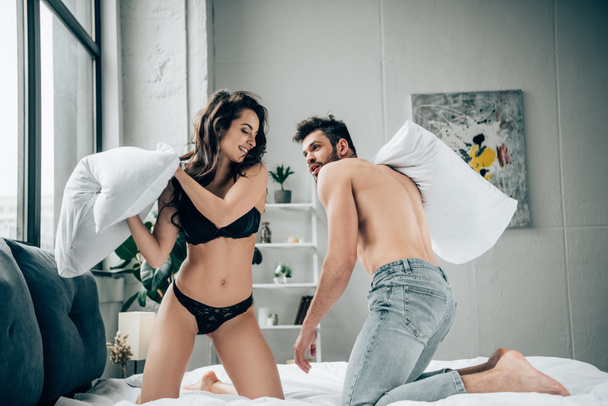cheerful brunette woman in lace underwear having pillow fight with muscular boyfriend on bed  - Foto, Imagen