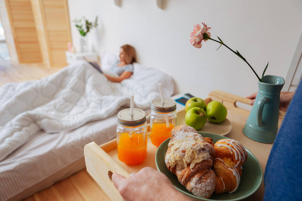 Wife lying in bed while husband bringing tray with croissants and fruits - Foto, Imagen