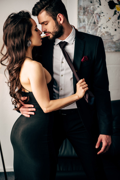 attractive woman in black dress holding tie of passionate man standing in suit  - Photo, Image