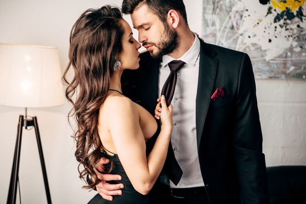 attractive girl in black dress holding tie of passionate man standing in suit  - Photo, Image