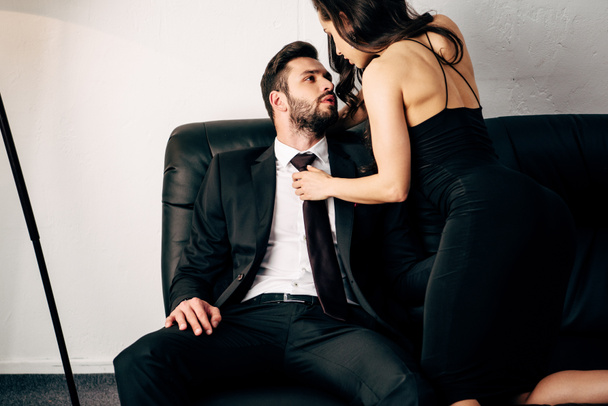 brunette girl in black dress holding tie of passionate man in suit sitting on sofa  - Фото, зображення
