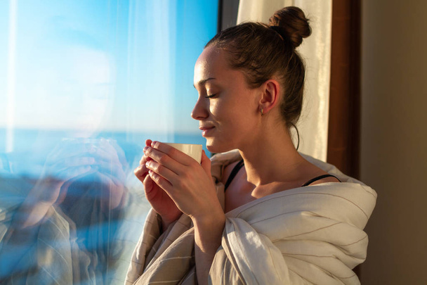 Kaunis nuori nainen kääritty huopa seisoo lähellä ikkunaa ja nauttia ensimmäinen aamu kahvia auringonpaisteessa. Varhainen herääminen ja uuden päivän alku
  - Valokuva, kuva