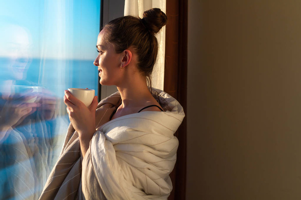 Csinos, fiatal nő burkolózva egy takarót ablak közelében, és élvezi az első reggeli kávé a napsütés. Egy korai ébredés és a kezdete egy új nap  - Fotó, kép