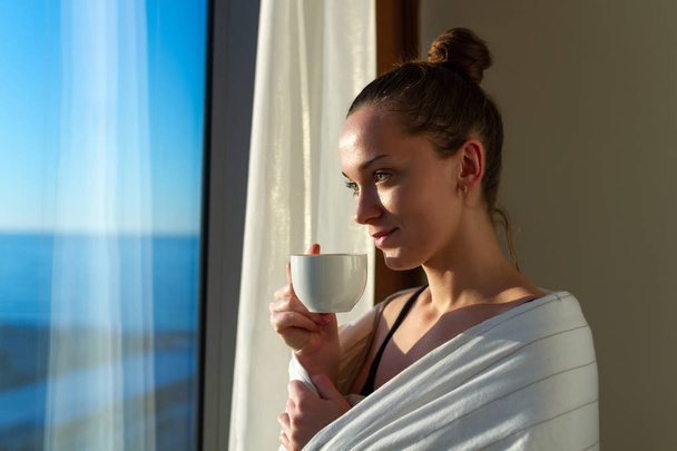 Pretty young woman wrapped in a blanket is standing near window and enjoying first morning coffee on sunshine. An early waking and the beginning of a new day  - Fotoğraf, Görsel