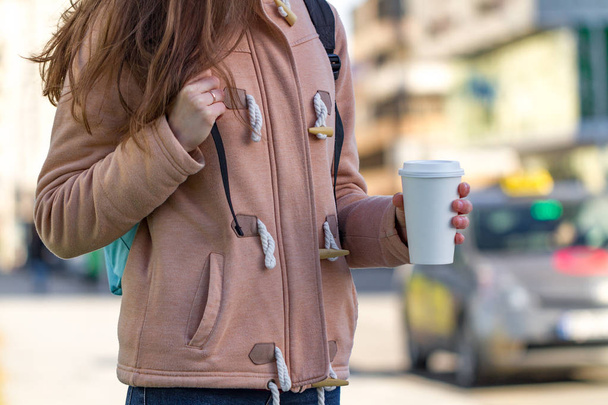 Eine junge Frau in lässiger Kleidung hält eine Kaffeetasse aus Papier in der Hand und genießt den morgendlichen Spaziergang in der Stadt an einem sonnigen Tag. Kaffee weg und weg  - Foto, Bild