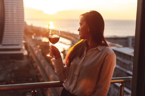 Young woman is relaxing and drinking a glass of red wine on the balcony at sunset in the evening. - Φωτογραφία, εικόνα