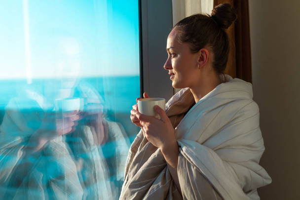 Pretty young woman wrapped in a blanket is standing near window and enjoying first morning coffee on sunshine. An early waking and the beginning of a new day  - 写真・画像