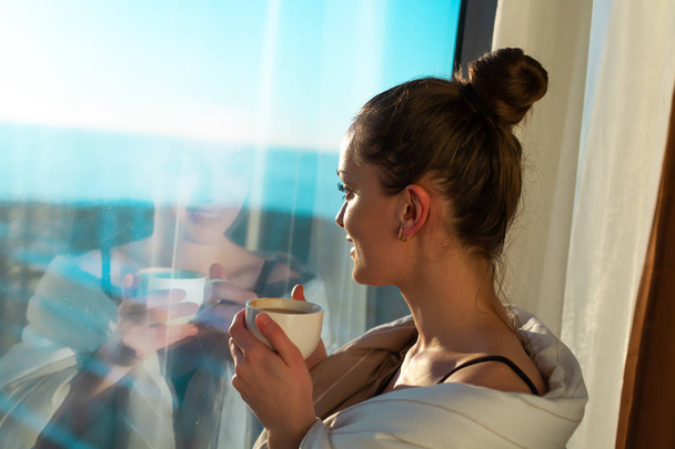 hübsche junge Frau in eine Decke gehüllt steht am Fenster und genießt den ersten Morgenkaffee bei Sonnenschein. ein frühes Erwachen und der Beginn eines neuen Tages  - Foto, Bild