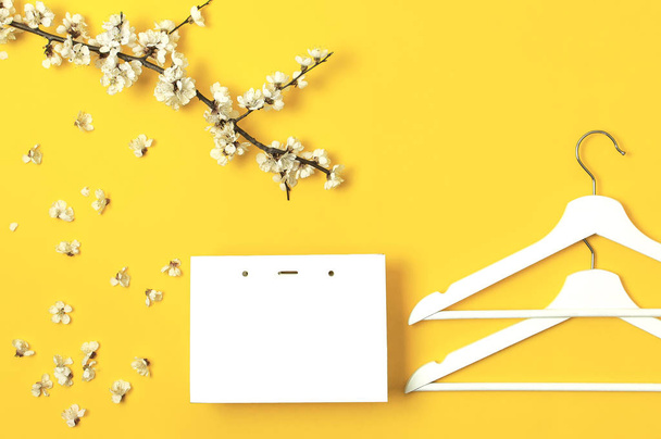 Witte houten hangers, voorjaar bloeiende tak, witte geschenk tas op gele achtergrond. Lente verkoop concept discount winkel winkels leeg hanger. Creatieve fashion banner. Plat lag bovenaanzicht kopie ruimte. - Foto, afbeelding