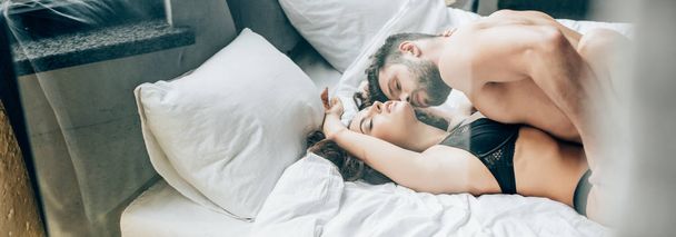 Panoramaaufnahme eines bärtigen Mannes ohne Hemd, der leidenschaftliche brünette Frau auf dem Bett küsst  - Foto, Bild