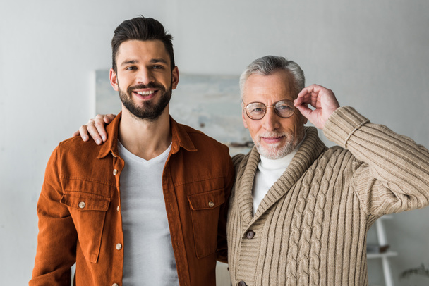 handsome man smiling while standing with cheerful senior father touching glasses  - Photo, Image