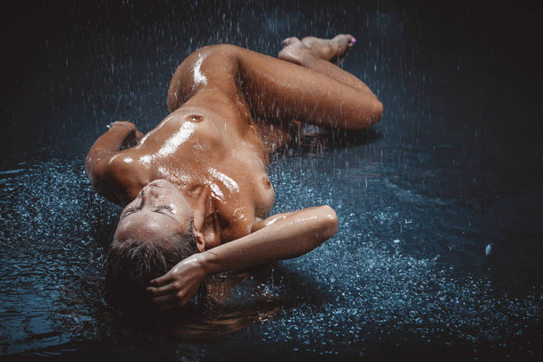 beautiful nude young woman lying on black floor under rain - 写真・画像