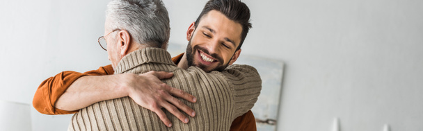 πανοραμικό στιγμιότυπο της ευτυχισμένη γενειοφόρος άνδρας χαμογελώντας ενώ αγκαλιάζει γέροντα πατέρα στο σπίτι  - Φωτογραφία, εικόνα