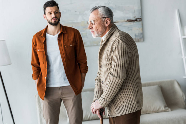 handsome bearded son standing with hands in pockets and looking at sad senior father in glasses - Fotoğraf, Görsel