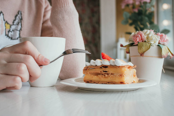 Classic french dessert millefeuille on a wooden board. Napoleon cake on tray or table with mint and a cup of coffee and flower decoration on the background. Girl or woman eating with fork In the local - Valokuva, kuva