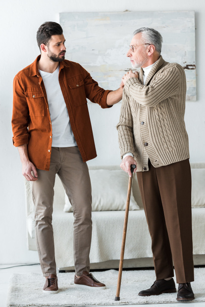 handsome bearded son putting hand on shoulder of sad senior father in glasses with walking stick - Photo, Image