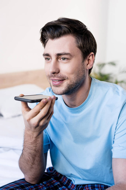 handsome smiling man talking on smartphone by speakerphone in bedroom - Фото, изображение
