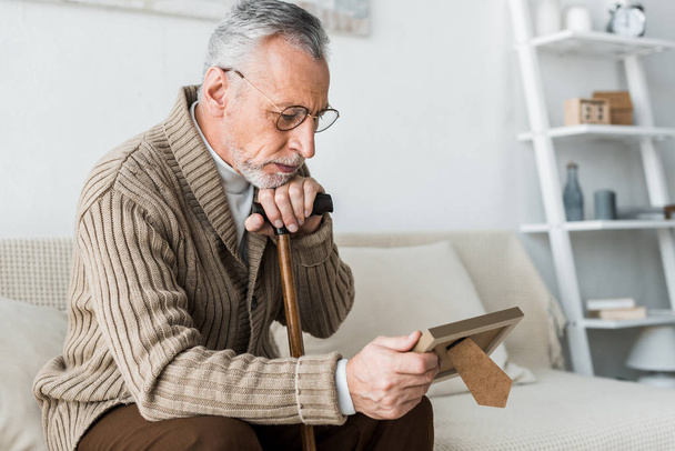 upset retired man looking at photo frame while holding walking stick - 写真・画像