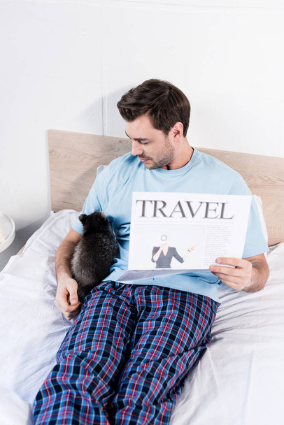 όμορφος άνθρωπος αγκαλιές στο κρεβάτι με αξιολάτρευτο ρακούν και κρατώντας εφημερίδα ταξιδιωτικό - Φωτογραφία, εικόνα