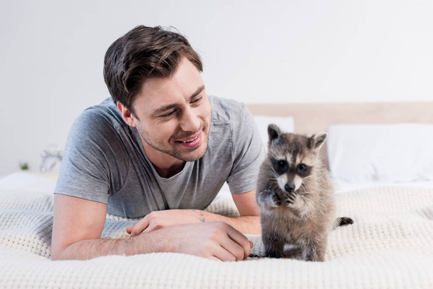 handsome cheerful man resting on bedding with funny raccoon - Zdjęcie, obraz