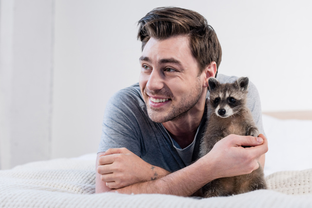 cheerful man cuddling with adorable raccoon on bed at home - Foto, afbeelding