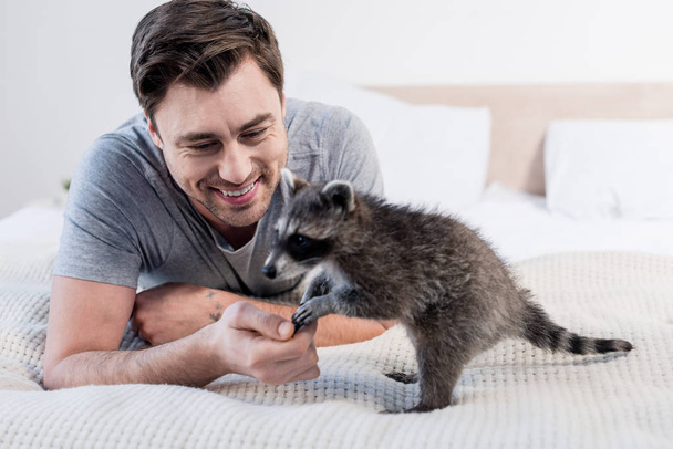 handsome man playing with funny raccoon on bedding at home - Foto, Imagen