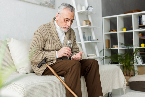retired man holding pill and glass of water while sitting on sofa near walking cane  - Fotó, kép