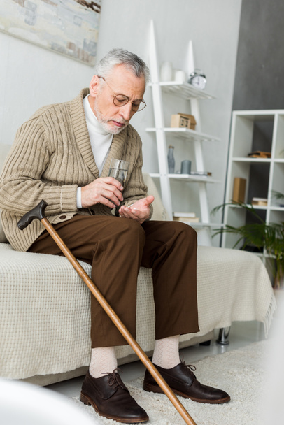 senior man holding pill and glass of water while sitting on sofa near walking cane  - Фото, изображение