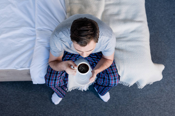 vue de dessus de l'homme en pyjama tenant une tasse de café tout en étant assis sur la literie
 - Photo, image