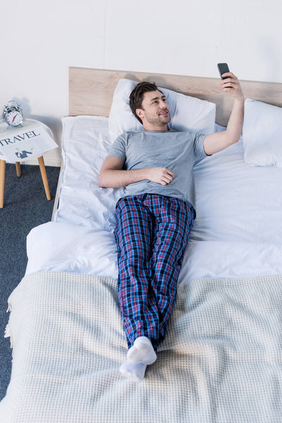 handsome man taking selfie with smartphone while resting on bed at home - Foto, Imagen