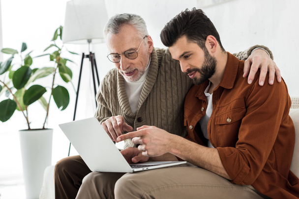 handsome bearded man pointing with finger at laptop while sitting with senior father  - Foto, Bild