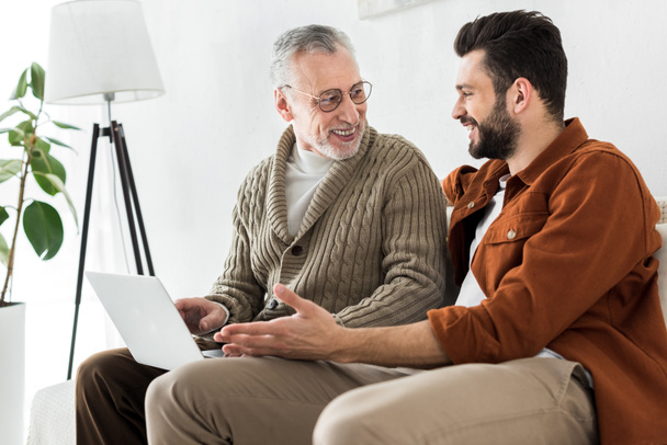 happy bearded man gesturing while sitting with senior father and looking at laptop  - Photo, image